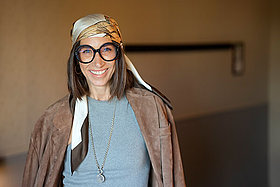 Frau mit langen dunklen Haaren, dick umrandeter Brille und stylischem Kopftuch. 