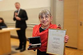 Dr. med. Christina Smaczny im November 2023 bei der Verleihung der Theodor Stern-Medaille der Universitätsmedizin Frankfurt. Sie hält eine Urkunde in die Kamera und trägte einen roten Schal. Im Hintergrund ist verschwommen ein anderer Mensch zu sehen. 