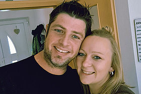 Selfie von Stefan (links, dunkle kurze Haare, schwarzer Pullover) und Franziska (rechts, lange blonde Haare, zu einem Zopf gebunden), beide lachen leicht in die Kamera.