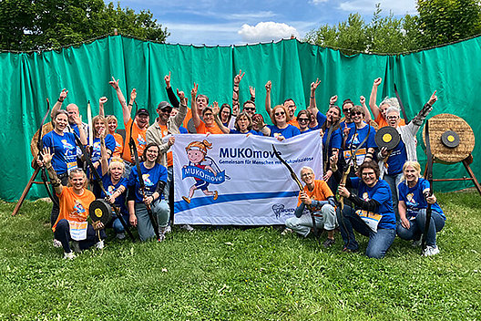 Betriebsausflug des Mukoviszidose e.V. zum MUKOmove 2025 Eine Gruppe von etwa 35 Menschen steht vor einer grünen Plane, daneben sieht man Zielscheiben. Einige Menschen halten Bogen in der Hand. Außerdem halten sie ein Banner mit der Aufschrift MUKOmove in die Kamera. Die Menschen tragen blaue oder orangene T-Shirts.
