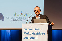 Mann auf Podium, an dem ein Banner mit der Aufschrift "Gemeinsam Mukoviszidose besiegen" hängt: Prof. Dr. Jürg Barben vom Ostschweizer Kinderspital St. Gallen
