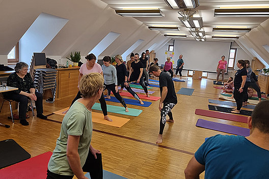 Seminarraum mit mehreren Menschen auf Yogamatten, die gerade eine Übung durchführen und dadurch ein bisschen in die Knie gehen. 