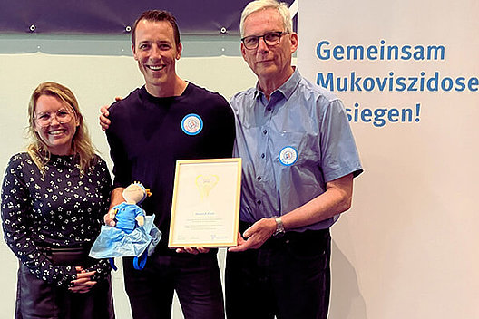 Eine jüngere Frau (links) und ein älterer Mann (rechts) übergeben eine Urkunde und eine Stoffpuppe mit Handball-Kleidung an einen Mann in dunkler Kleidung in der Mitte.