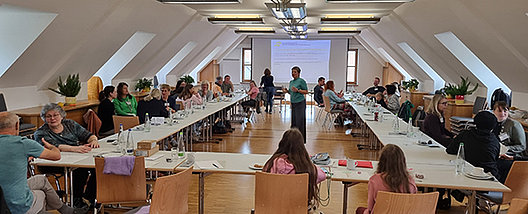 Seminarraum mit Tischen in U-Form, an denen Menschen sitzen und zum Referenten nach vorne blicken. 