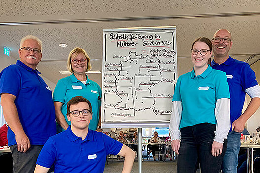 Drei Männer (in blauen Shirts) und zwei Frauen (in türkisen Shirts) stehen vor einer gemalten Deutschlandkarte auf einem Flipchart, auf dem "Selbsthilfe-Tagung in Münster" zu lesen ist. 