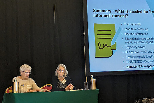 Zwei Frauen auf Podium vor schwarzer Wand, dazu Projektion auf Leinwand: "What is needed for informed consent?"