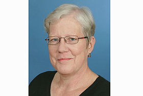 Porträt von Birgit: Graue Haare, schwarzer Pulli, blauer Hintergrund. Birgit hat Mukoviszidose und wird 2024 70 Jahre alt.