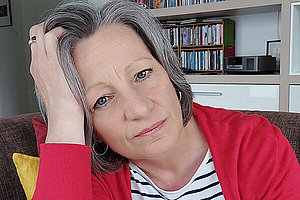 Porträt von Simona, graue Haare, rote Strickjacke, ca. 55 Jahre alt. Sie stützt ihren Kopf mit ihrem Arm. 