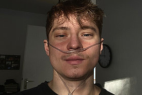 Jonas (junger Mann, dunkele Haare, dunkle Kleidung) hat eine Sauerstoffbrille auf. 