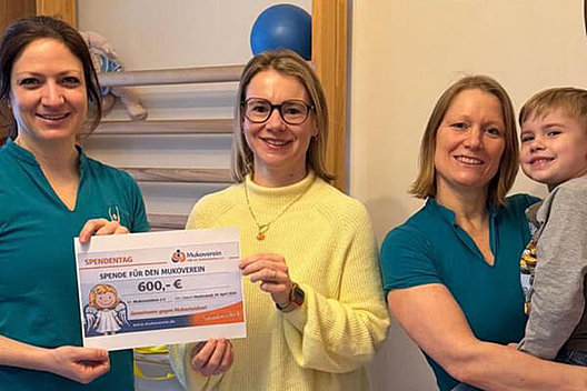 Drei Frauen und ein Kind vor einer Sprossenwand, zwei der Frauen halten einen Spendenscheck für den Mukoviszidose e.V. über 600 Euro in den Händen. 