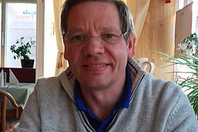 Mann, ca. 59 Jahre alt, kurze Haare, Brille, sitzt in einem Raum.