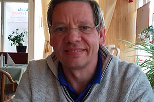 Mann, ca. 59 Jahre alt, kurze Haare, Brille, sitzt in einem Raum.
