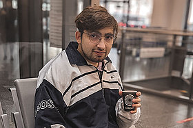 Junger Mann mit dunklen Haaren, sitzt mit einem Papp-Kaffebecher in der Hand in einer Eingangshalle. Üzeyir hat Mukoviszidose und setzt seine Hoffnung in die Forschung