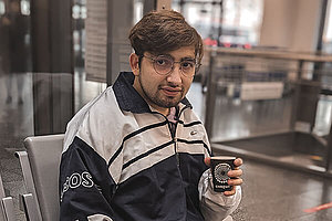 Junger Mann mit dunklen Haaren, sitzt mit einem Papp-Kaffebecher in der Hand in einer Eingangshalle. Üzeyir hat Mukoviszidose und setzt seine Hoffnung in die Forschung