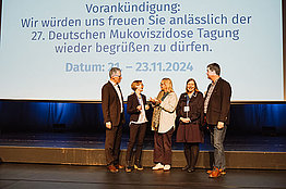 Stephan Kruip (links), Miriam Schlangen (Mitte) und Winfried Klümpen (rechts) bedanken sich bei den Tagungsleiterinnen Prof . Dr . Anna-Maria Dittrich (2.v.l) und Dr . Susanne Nährig (2.v.r.). Im Hintergrund sieht man den Termin der nächsten Tagung. 