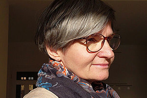 Porträt einer etwa 50-jährigen Frau, sie schaut zur Seite, hat kürze graumelierte Haare und eine Brille