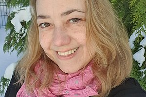 Frau mit blonden Haaren und rosa Schal lächelt in die Kamera, man sieht eine Tanne mit Schnee im Hintergrund. 