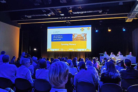 Opening Plenary bei der europäischen CF-Konferenz Konferenzsaal, man sieht viele Hinterköpfe, vorne ist eine beleuchtete Bühne, eine Folie ist an die Wand geworfen, auf der "Opening Plenary" steht.