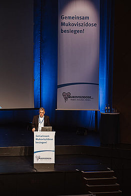 In klein sieht man einen Mann am Podium stehen, im Hintergrund sieht man ein großes Banner des Mukoviszidose e.V.: Stephan Kruip, Vorstandsvorsitzender des Mukoviszidose e.V., eröffnet die Deutsche Mukoviszidose Tagung 2023.