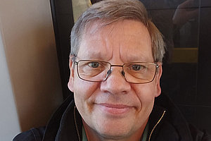 Porträt von Thomas: Er sitzt im Zug, hat anscheinend ein Selfie gemacht. Dunkelblonde Haare, Brille, Thomas lacht. Er ist rund 59 Jahre alt. 