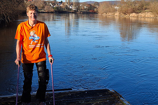 Frau mit Krücken steht vor einem See. Sie trägt ein MUKOmove-T-Shirt. 