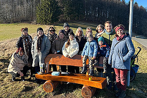 Foto einer Gruppe von Menschen in Winterkleidung vor einer Holzbank auf einer Wiese - im Hintergrund sieht man einen Wald. 