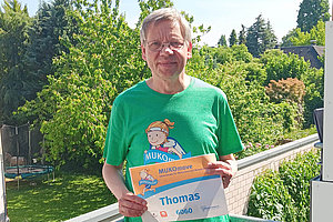 Mann, um die 60 Jahre alt, hält eine MUKOmove-Startnummer in der Hand, steht vermutlich auf einem Balkon. 