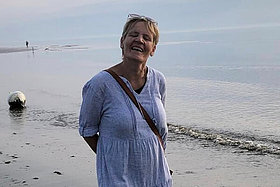 Eine ältere Frau mit blauem Kleid steht am Strand. 
