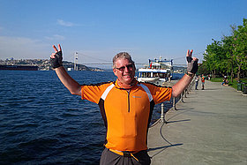 Mann in orangener Fahrradkleidung steht vor einem Meer / Fluss (vermutlich der Bosporus), reckt beide Arme in die Höhe und im Hintergrund sieht man eine sehr große Brücke. 
