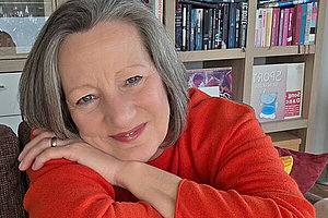 Porträt einer Frau um die 60 Jahre - roter Pullover, lächelt, der Kopf ist auf die Hände gelegt. Im Hintergrund steht ein Bücherregal. 