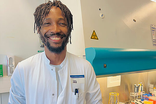 Porträt von Dr. Andrew Tony-Odigie in seinem Labor, er trägt einen Laborkittel und lächelt in die Kamera. 