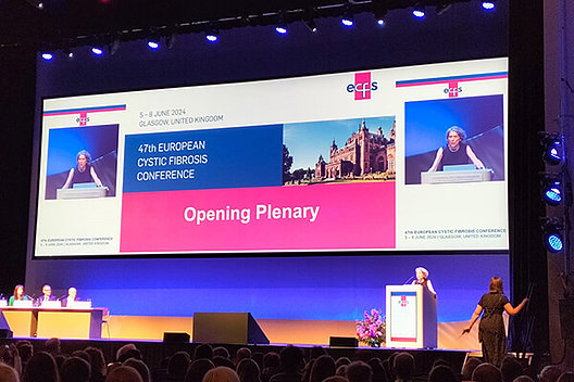 Blick auf die Bühne, auf der gerade ein Vortrag gehalten wird. Eine große Beamer-Projektion ist zu sehen, auf der man "Opening Plenary" lesen kann. Eröffnungsvortrag der Europäischen CF-Konferenz in Glasgow 2024. 