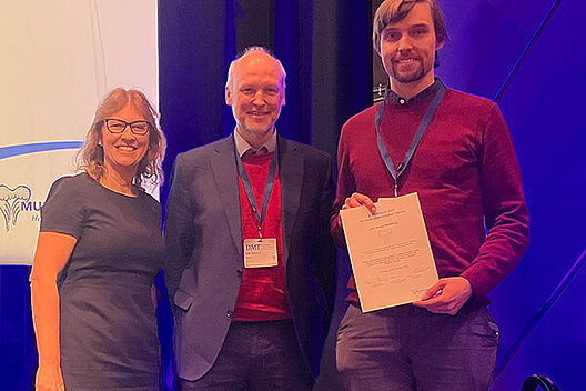 Zweiter Posterpreis an Dr. Julian Berges Drei Menschen (zwei Männer und eine Frau) posieren auf einer Bühne, einer hält eine Urkunde in der Hand.