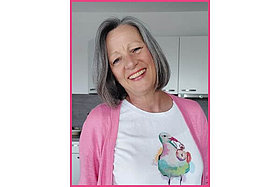 Porträt von Simona: Dunkelgrau-melierte Haare, rosa Strickjacke, etwa 60 Jahre alt, lächelt. 