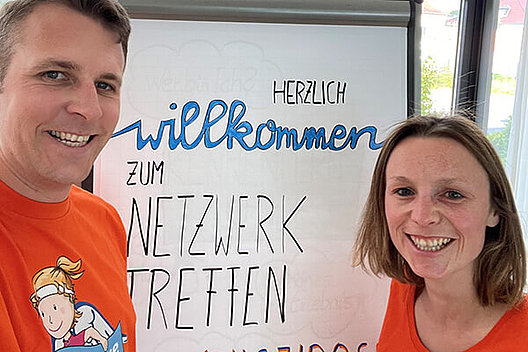 Erstes Netzwerktreffen für Menschen mit Mukoviszidose und ihre Angehörigen in der Region Fulda Ein Mann und eine Frau, beide tragen orangene T-Shirts, stehen vor einem Flip-Chart, auf dem "Netzwerktreffen" zu lesen ist.