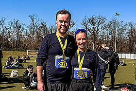 Ein Mann und eine Frau in Sportkleidung mit Medaillen um den Hals lachen bei blauem Himmel in die Kamera. 