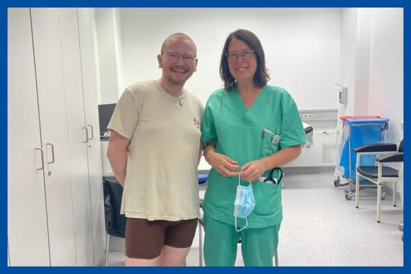 Junger Mann in kurzen Sachen, kurze rote Haare, steht neben einer Ärztin in grünem Arztkittel. Im Hintergrund sieht man ein Sprechzimmer. 