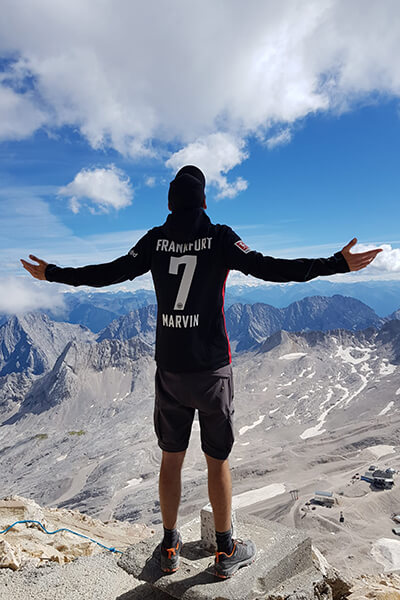 Mann im FC Frankfurt Trikot auf der Zugspitze bei Sonnenschein, steht mit dem Rücken zur Kamera, im Hintergrund sieht man die Berge. Marvin hat sich mit der Wanderung zur Zugspitze einen großen Traum erfüllen können. 