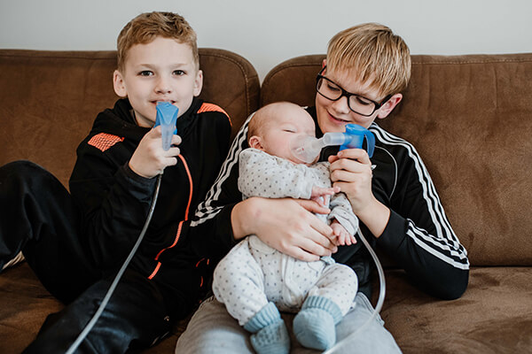 Drei Brüder, die zwei jüngeren inhalieren. Drei Jungen sitzen auf einem Sofa, der ältere hält seinen kleinen Bruder, ein Baby auf dem Schoß, und hilft ihm beim Inhalieren. Auch der mittlere Junge inhaliert.