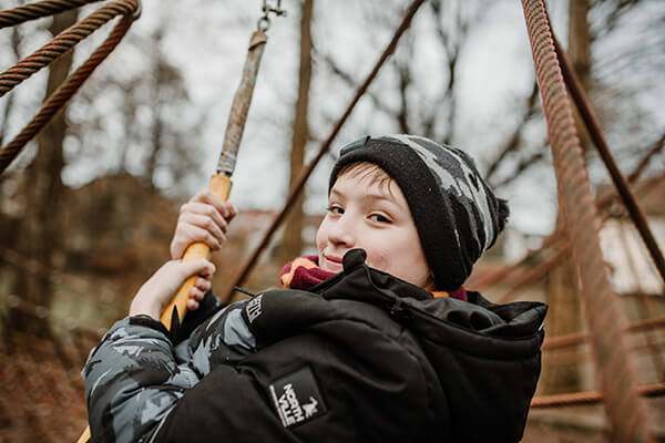 Junge in Winterkleidung auf dem Spielplatz, er scheint auf einer Schaukel oder einem ähnlichen Spielgerät zu hängen. 