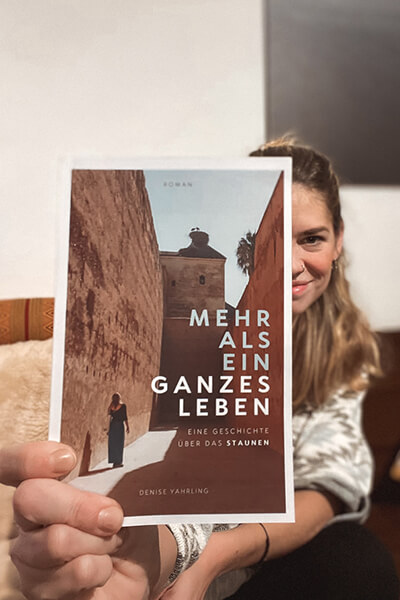 Denise hält ihr Buch "Mehr als ein ganzes Leben" in die Kamera. Auf dem Cover sieht man eine junge Frau in einer vermutlich marokkanischen Stadt neben hohen Mauern in der Sonne stehen. 