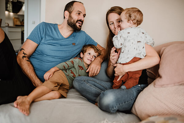 Eine Familie (Vater, Mutter und zwei kleine Kinder) sitzen auf einem Sofa. Sie lachen. 