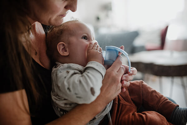 Ein Baby sitzt auf dem Schoß der Mutter und inhaliert mit einem Feuchtinhalator. 