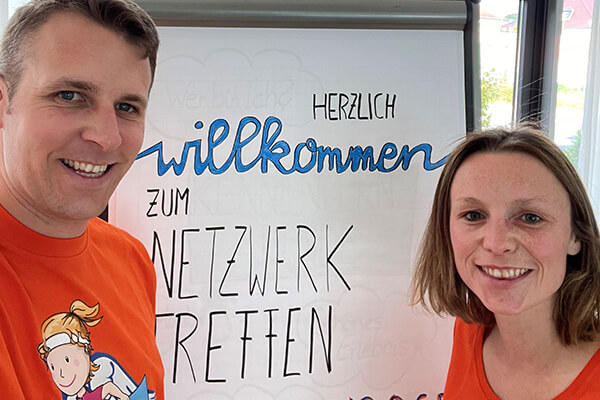Mann und Frau in orangenen T-Shirts stehen vor einem Flipchart, auf dem man "Herzlich Willkommen" und "Netzwerktreffen" lesen kann. 