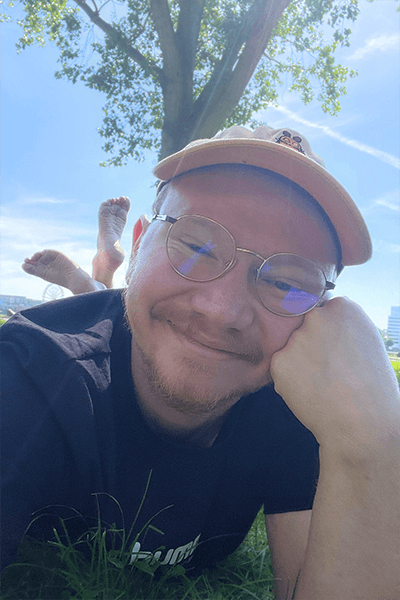 Jakob im Sommer im Park, er liegt auf einer Wiese, im Hintergrund sieht man einen Baum und blauen Himmel. 