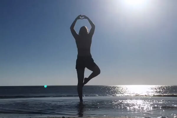 Sandra steht am Strand auf einem Bein, ihre Hände treffen sich über ihrem Kopf und formen ein Herz. 