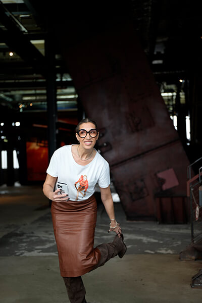 Frau mit weißem T-Shirt und braunem Lederrock hat ein Bein angehoben, steht wahrscheinlich irgendwo draußen, der Hintergrund ist aber nicht so gut zu erkennen. 
