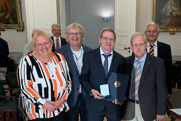 Festlich gekleidete Menschen, einer hält eine Medaille in der Hand. 