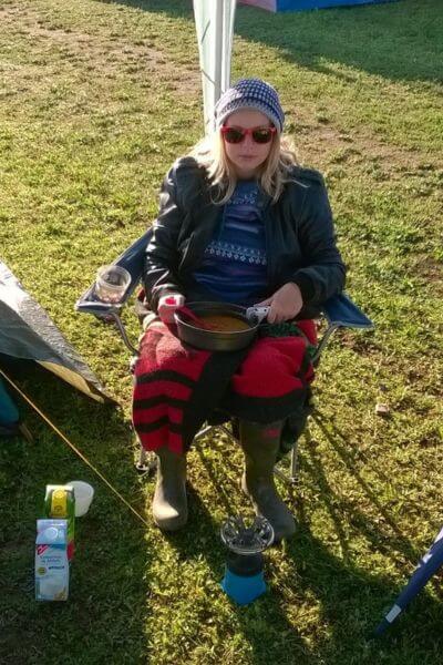 Zoë beim Festival: Da gibt es zum Frühstück schon mal Dosensuppe. Auf dem Bild sieht man eine Frau mit einer Suppenschale in der Hand eingewickelt in eine Decke auf einem Campingstuhl sitzen. 