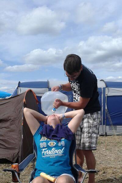 Haare waschen beim Festival. Eine Frau sitzt auf einem Campingstuhl mit dem Kopf weit zurück gelehnt, während ihr eine andere Person Wasser aus einem Kanister auf den Kopf schüttet. 